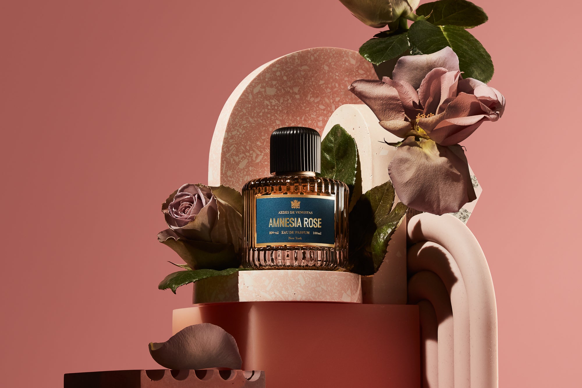 Parfumflakon Amnesia Rose von Aedes de Venustas mit geripptem Design und schwarzem Deckel auf geometrischen Podesten, dekoriert mit mauve Rosen und grünen Blättern vor rosa Hintergrund