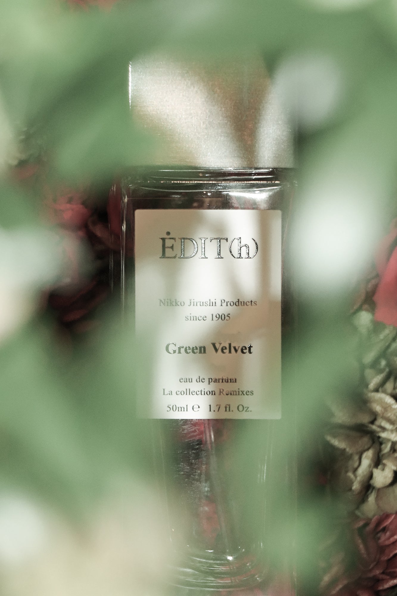 Nahaufnahme vom Parfumflakon Green Velvet von ÈDIT(h) mit cremefarbenem Etikett und goldfarbenem Verschluss hinter unscharfen grünen Blättern