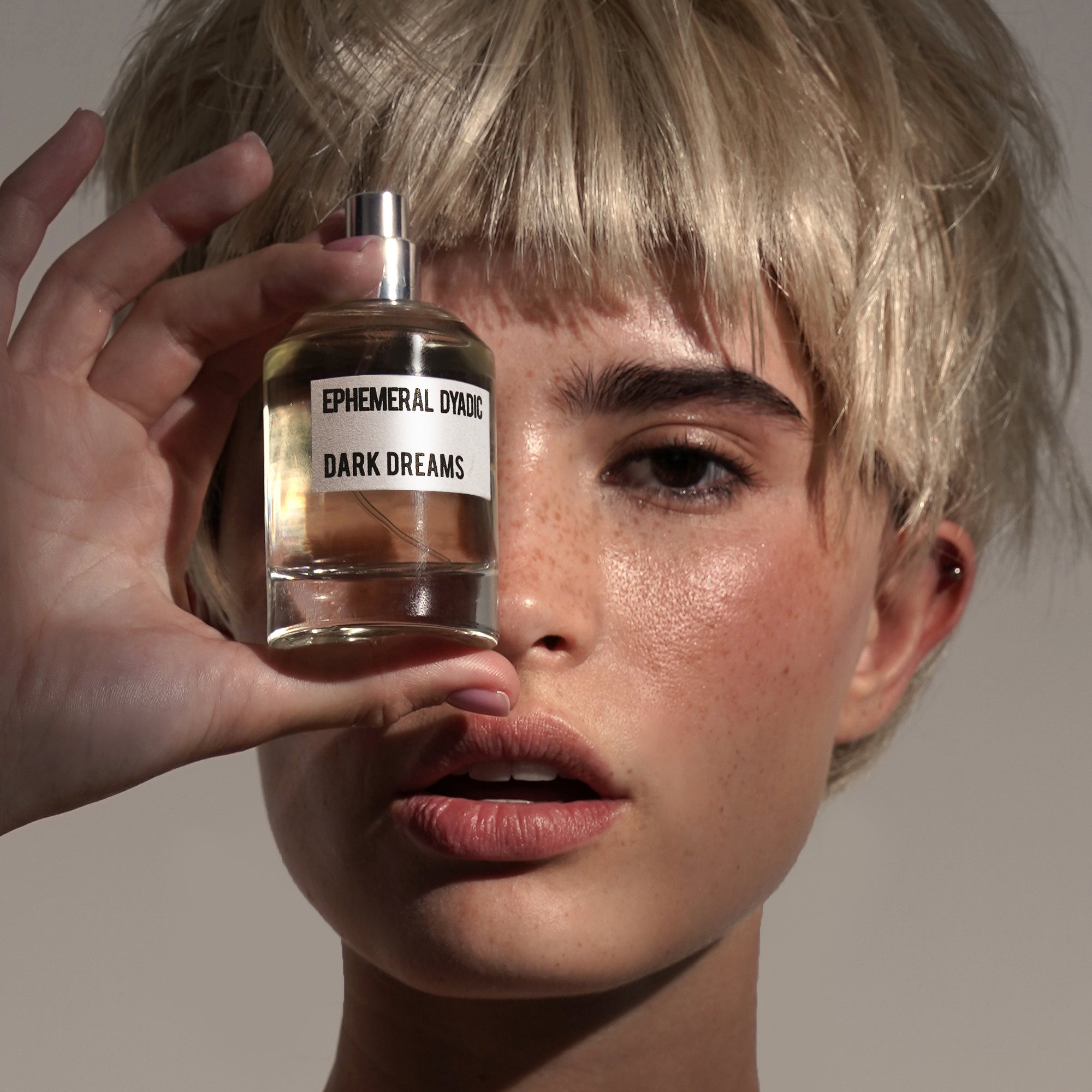 Frau mit blonden kurzen Haaren hält Parfumflakon Dark Dreams von Ephemeral Dyadic vor dem Gesicht vor neutralem Hintergrund