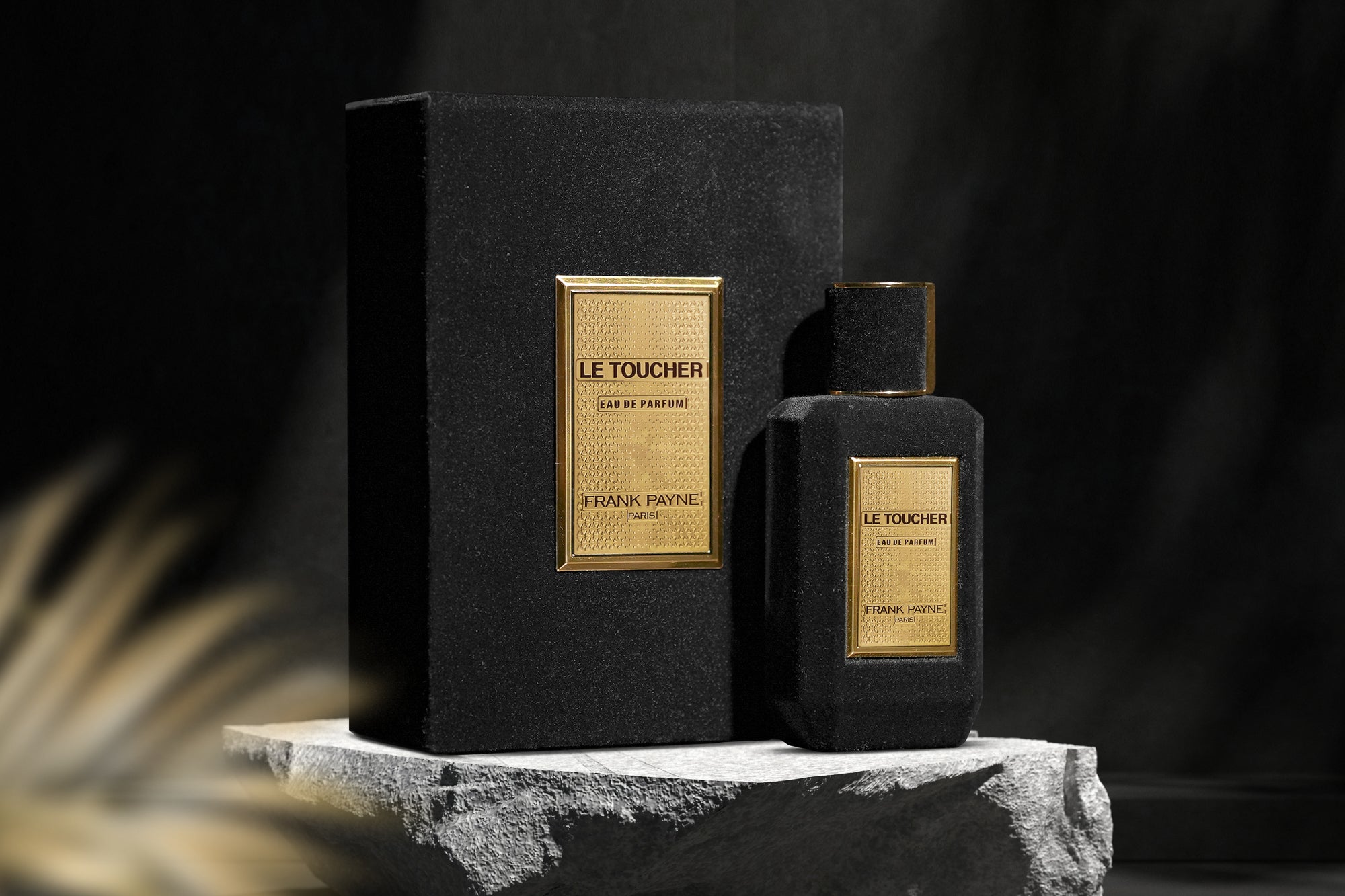 Schwarzer samtüberzogener Parfumflakon Le Toucher von Frank Payne mit goldenem Etikett und passender Verpackungsbox auf rauem Steinpodest vor dunklem Hintergrund