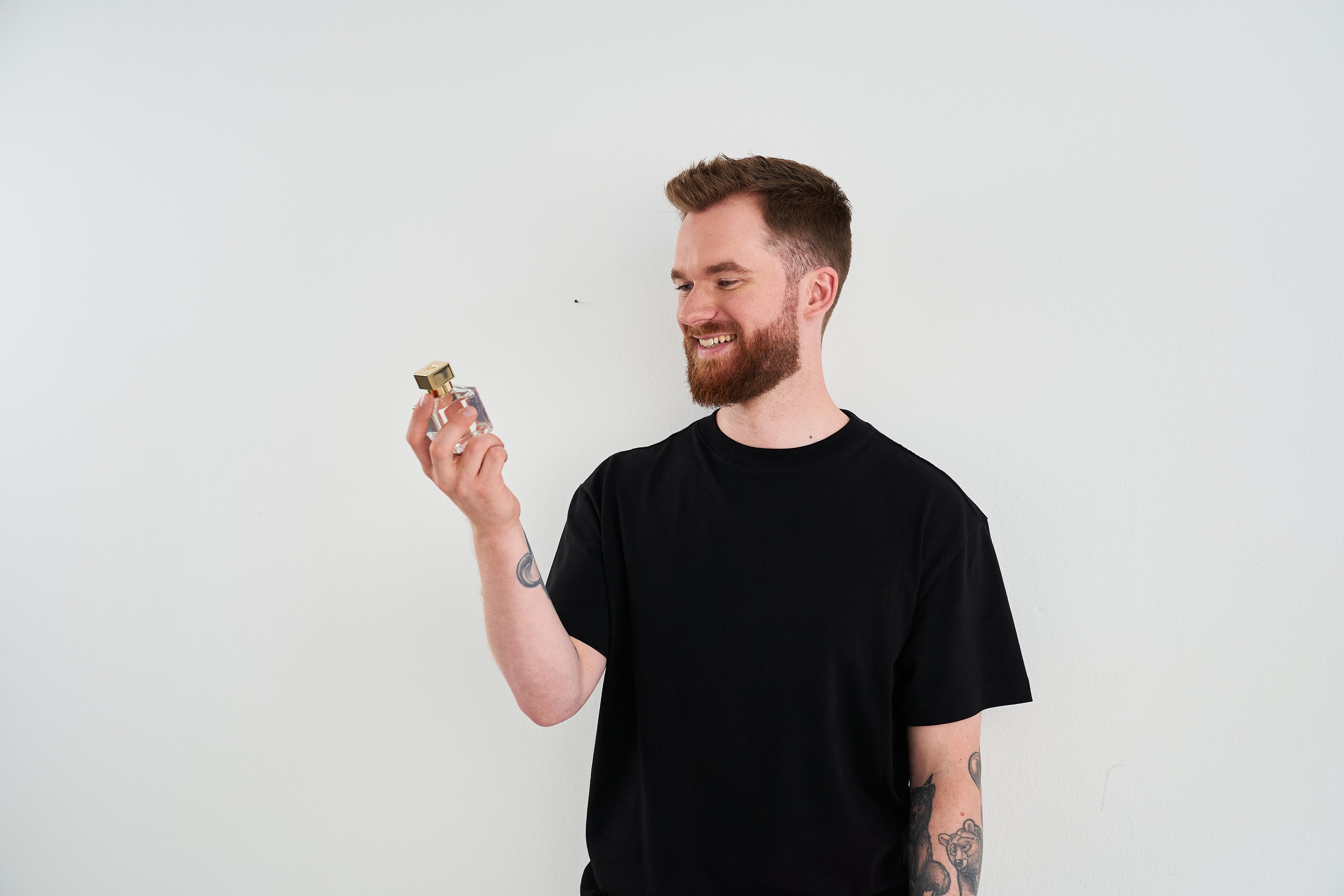 Porträt von Kai Porten in schwarzem T-Shirt, der lächelnd sein Parfum Oud 31 mit goldenem Deckel betrachtet, vor hellem Hintergrund