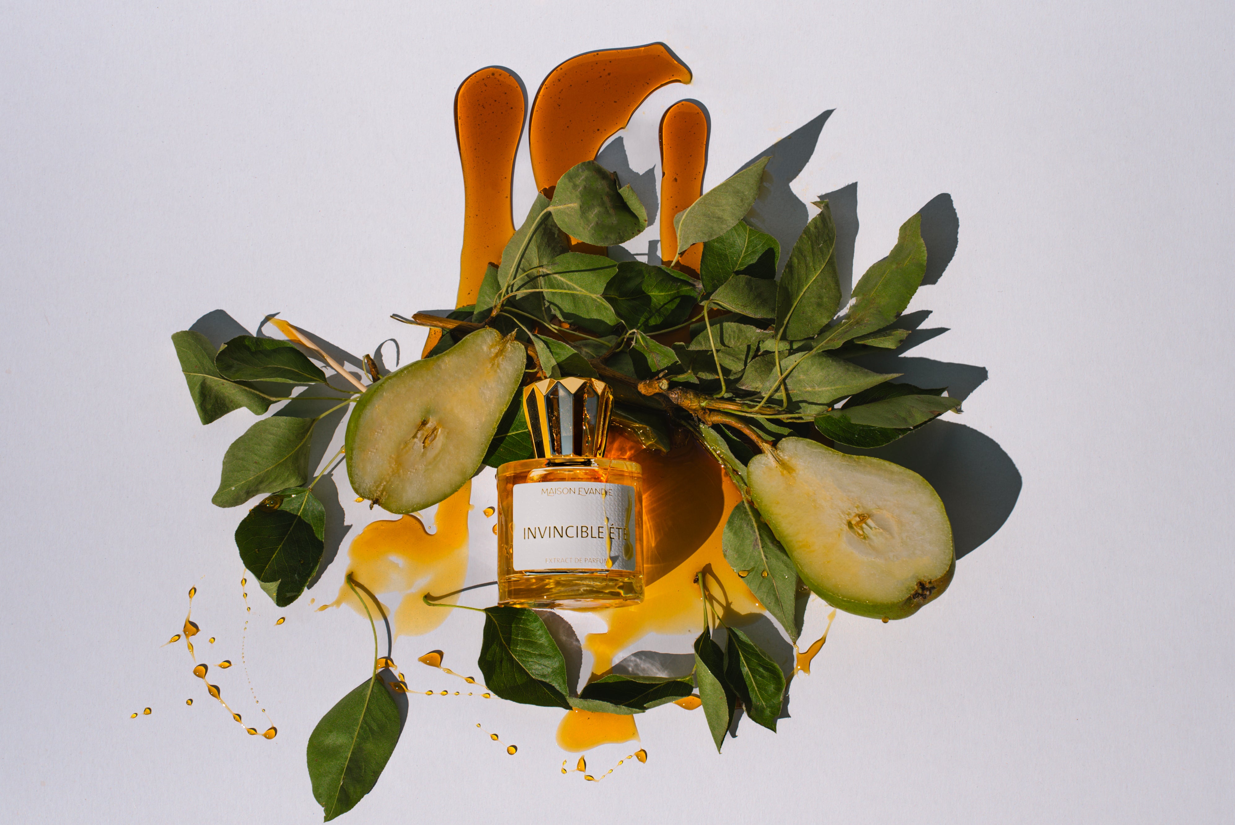 Parfumflakon Invincible Été von Maison Evandie mit goldener Flüssigkeit, arrangiert mit Birnen, grünen Blättern, orangefarbenen Elementen und goldenen Tropfen