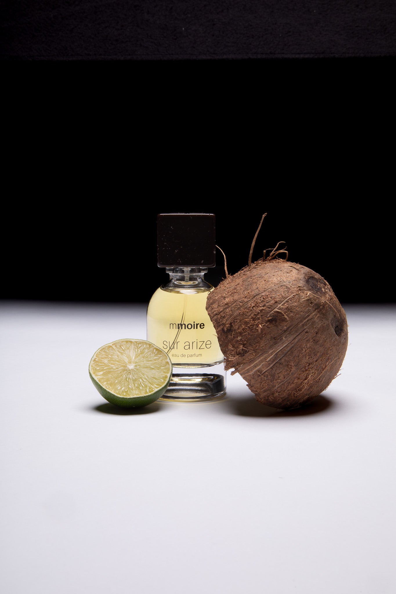 Parfumflakon Sur Arize von Mmoire mit schwarzem Verschluss, arrangiert mit halbierter Limette und Kokosnuss auf weißem Untergrund vor schwarzem Hintergrund
