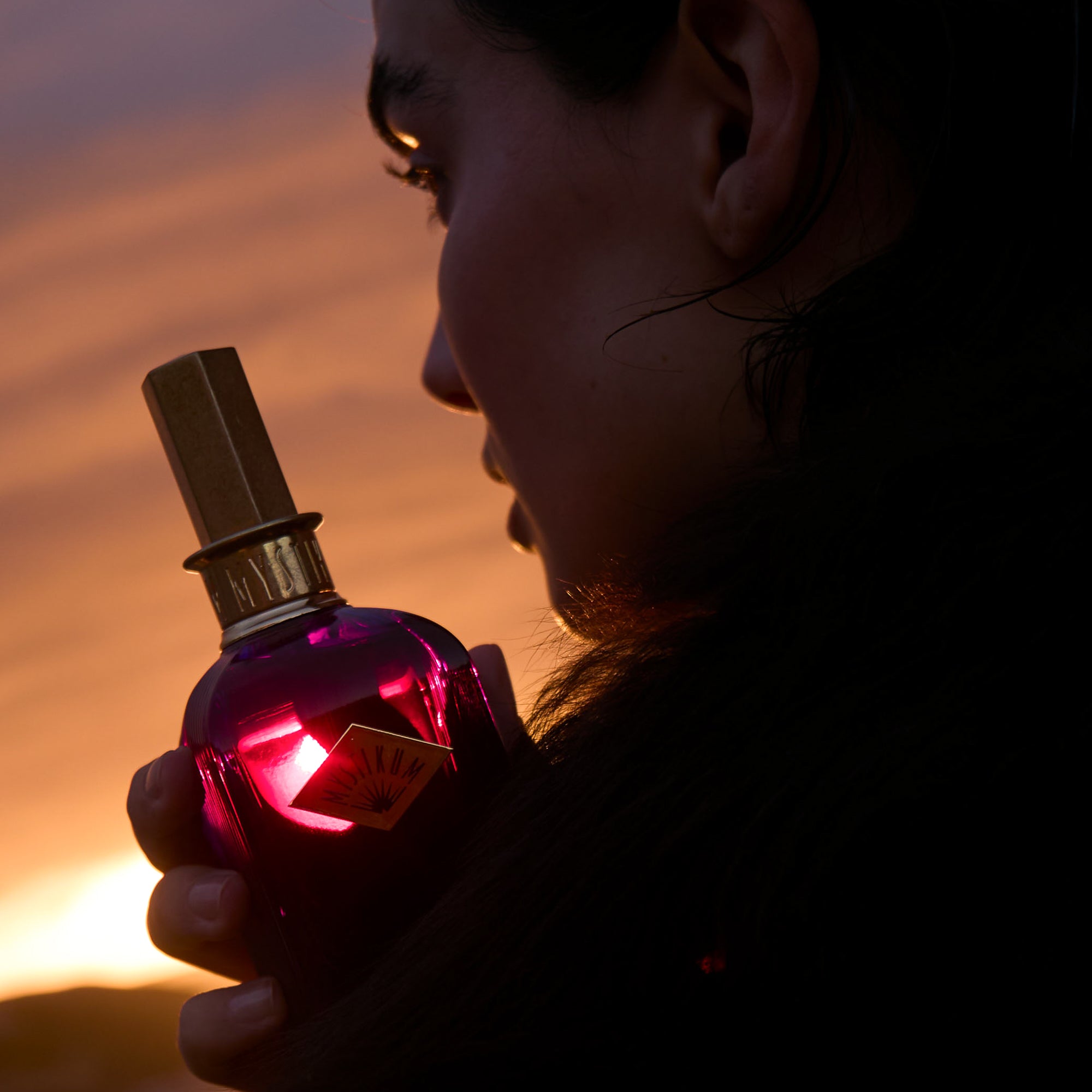 Frau hält rotfarbenen Parfumflakon The Empress Kiss von Mystikum mit facettiertem Glas und messingfarbenem Verschluss im Sonnenuntergangslicht