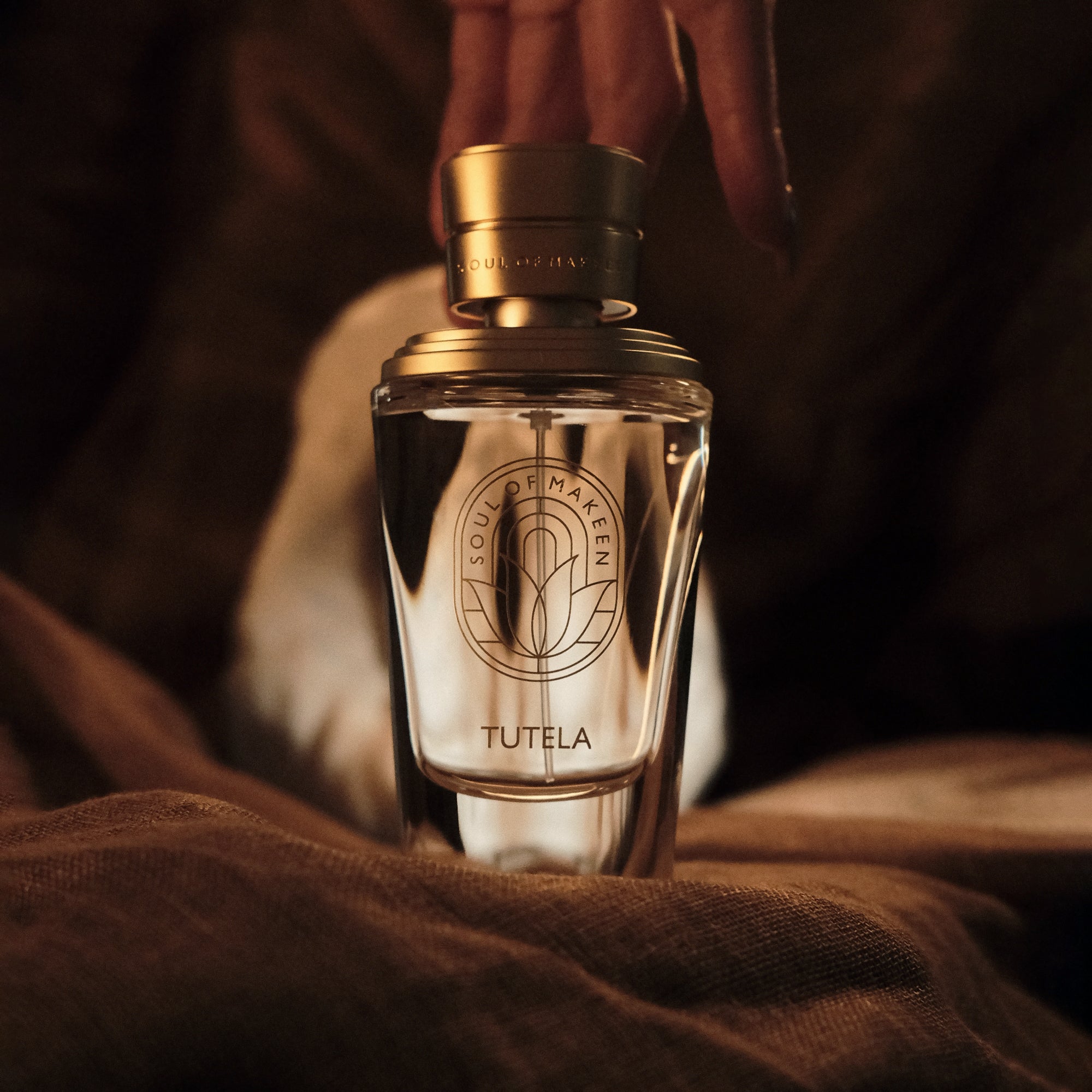 Parfumflakon Tutela von Soul of Makeen mit goldfarbenem Verschluss und geprägtem Logo auf braunem Stoff in warmer Beleuchtung