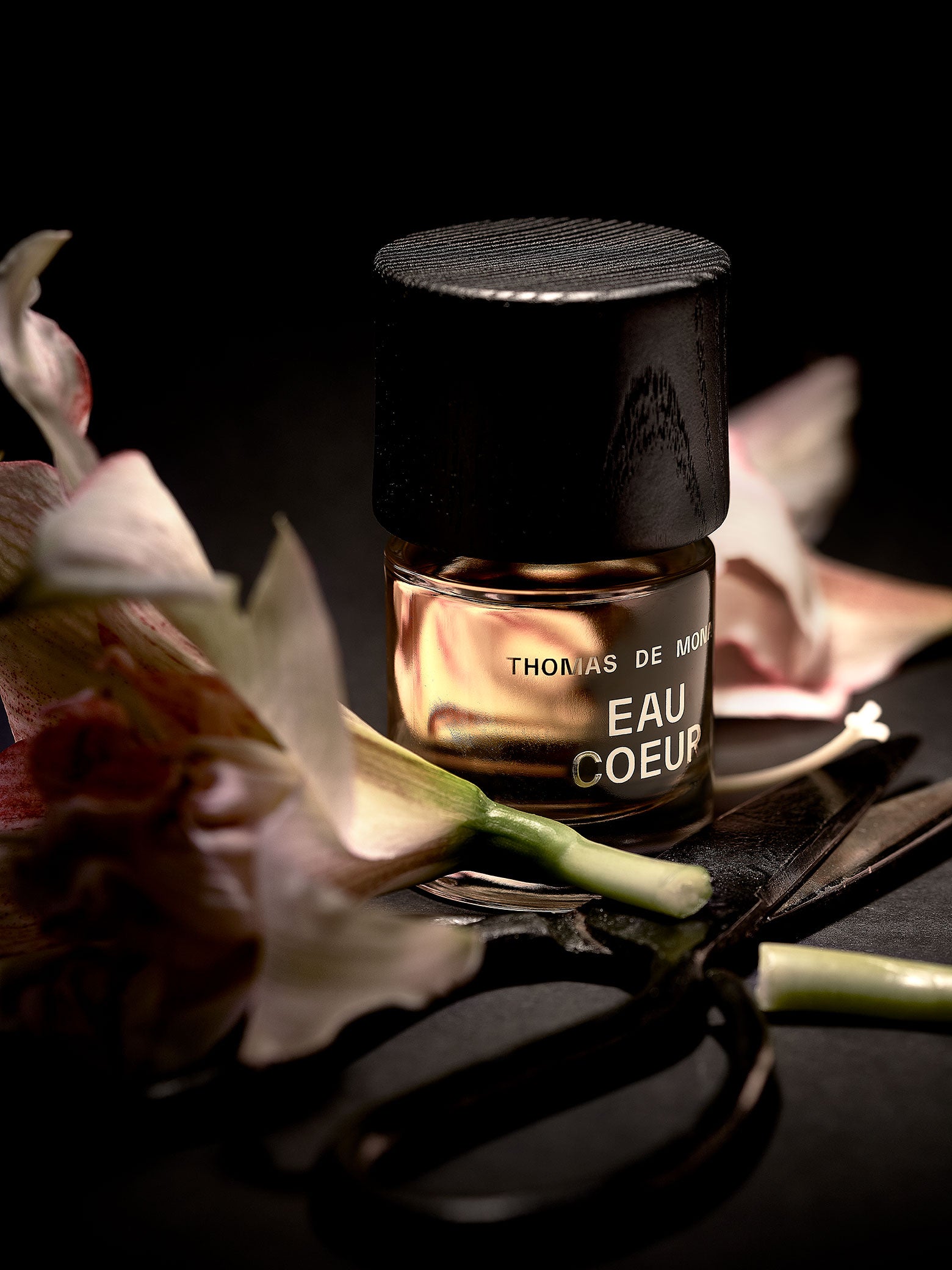 Parfumflakon Eau Coeur mit goldfarbig erscheinender Flüssigkeit und Holzverschluss, arrangiert mit Magnolienblüten und schwarzem Stoff