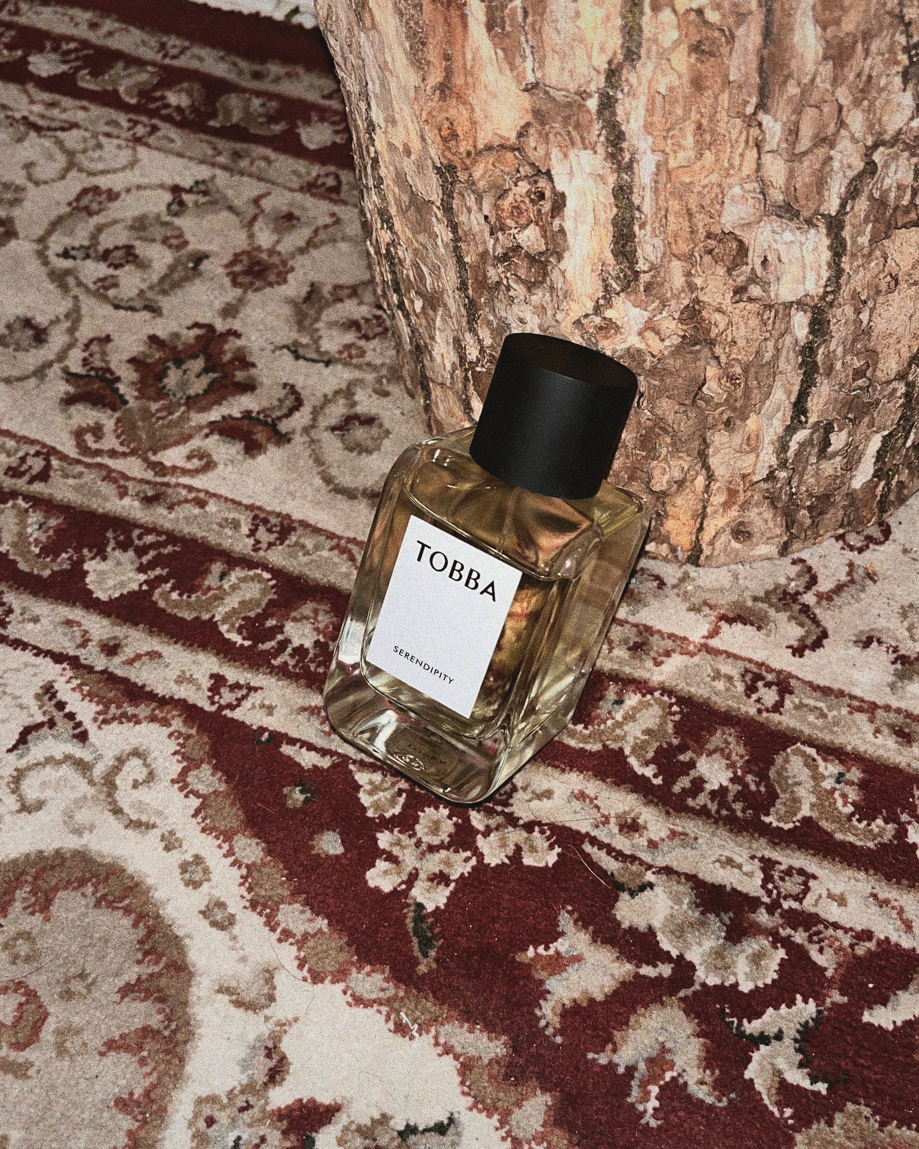 Parfumflakon Serendipity von Tobba mit schwarzem Verschluss auf beige-bordeauxrotem antiken Teppich vor Baumrinde