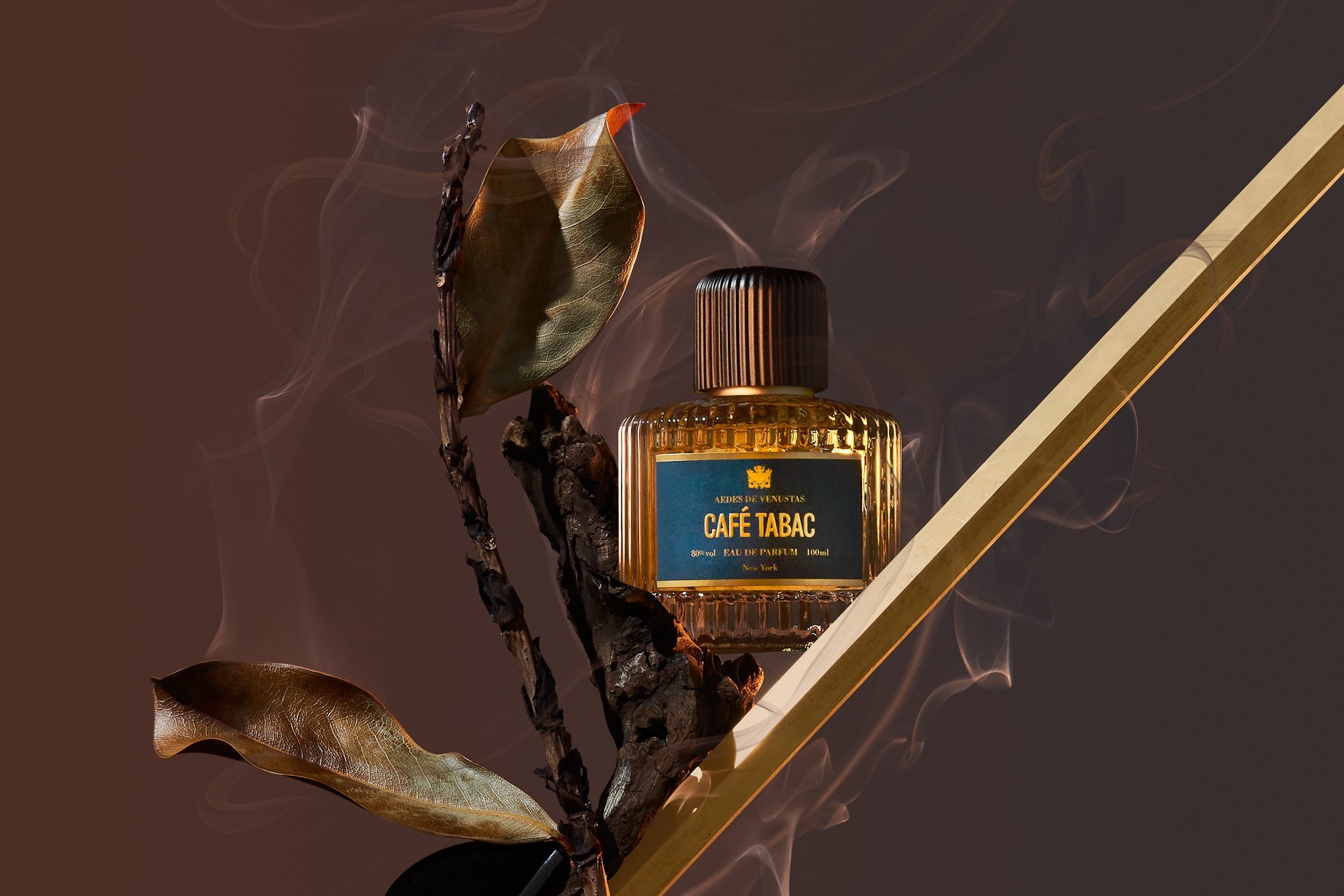 Parfumflakon Café Tabac von Aedes de Venustas mit geripptem Deckel auf dunklem Hintergrund mit Rauch, dekoriert mit verwelkten Blättern und goldenem Stab