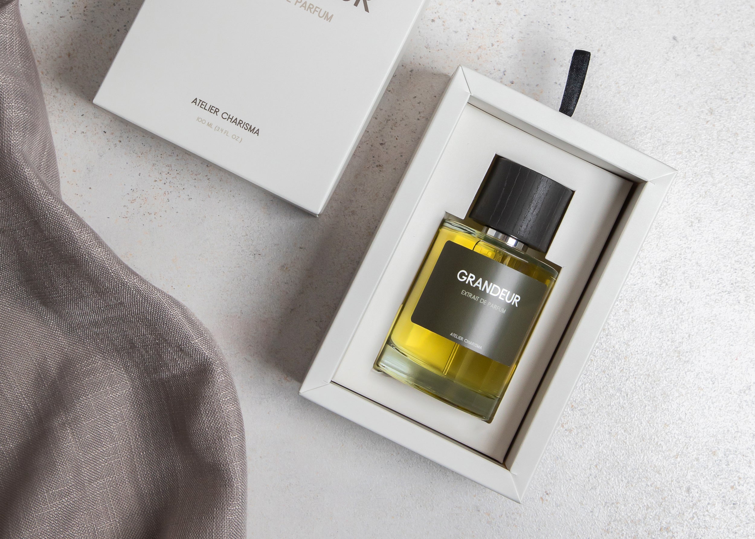 Parfumflakon Grandeur von Atelier Charisma mit goldener Flüssigkeit und schwarzem Deckel in weißer Verpackung mit grauem Tuch