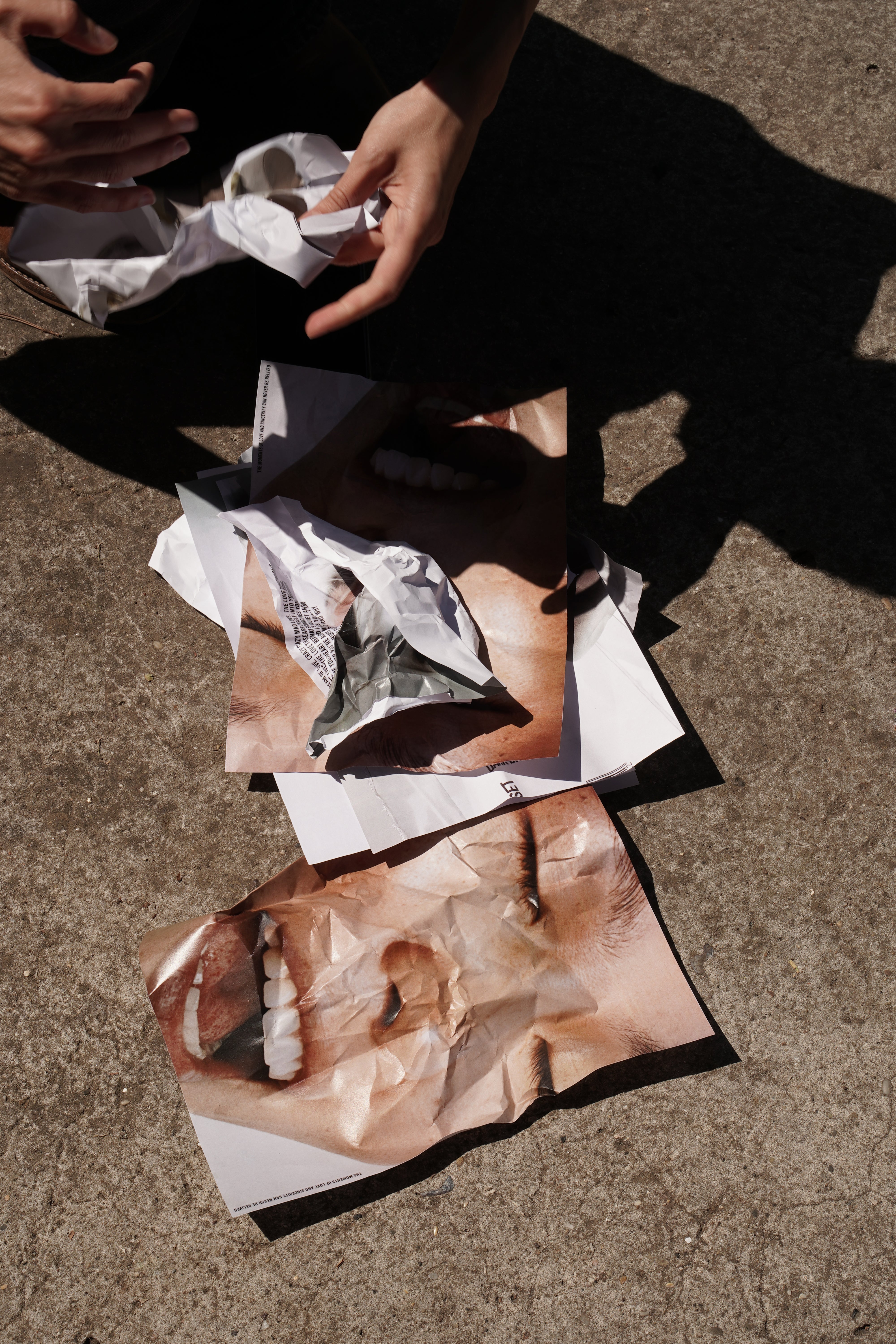 Vogelperspektive einer Person in Schwarz, die auf Beton sitzt und künstlerische Fotografien mit Körperdetails und zerknittertem Papier arrangiert