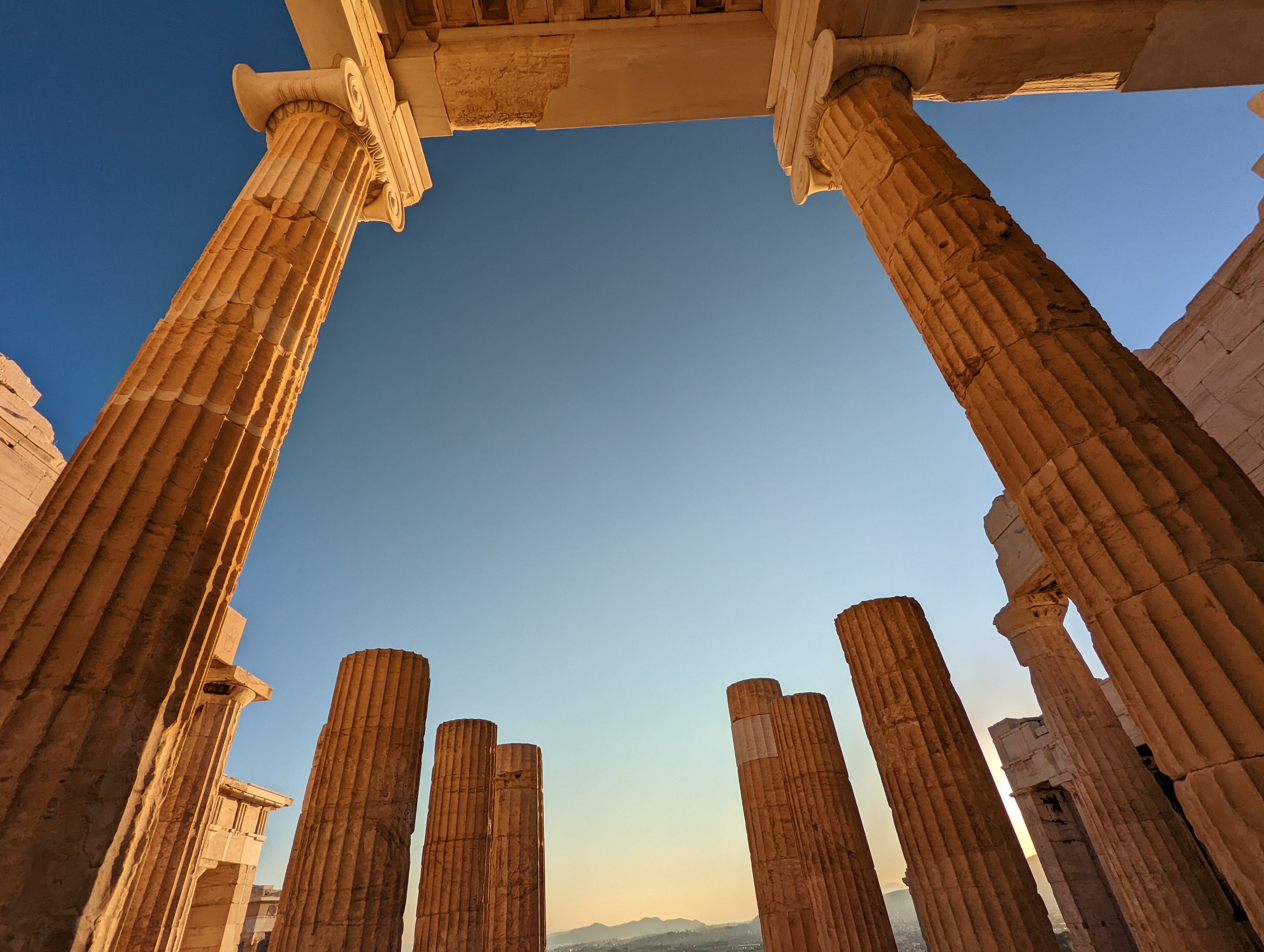 Blick nach oben durch monumentale dorische Säulen eines antiken griechischen Tempels vor blauem Himmel in goldenem Licht