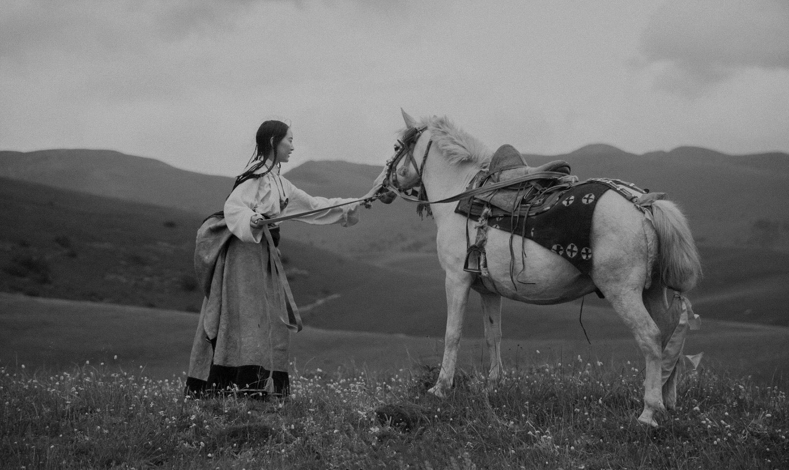 Schwarz-weiß Aufnahme einer Frau in traditioneller asiatischer Kleidung, die ein geschmücktes Pferd durch eine Berglandschaft führt
