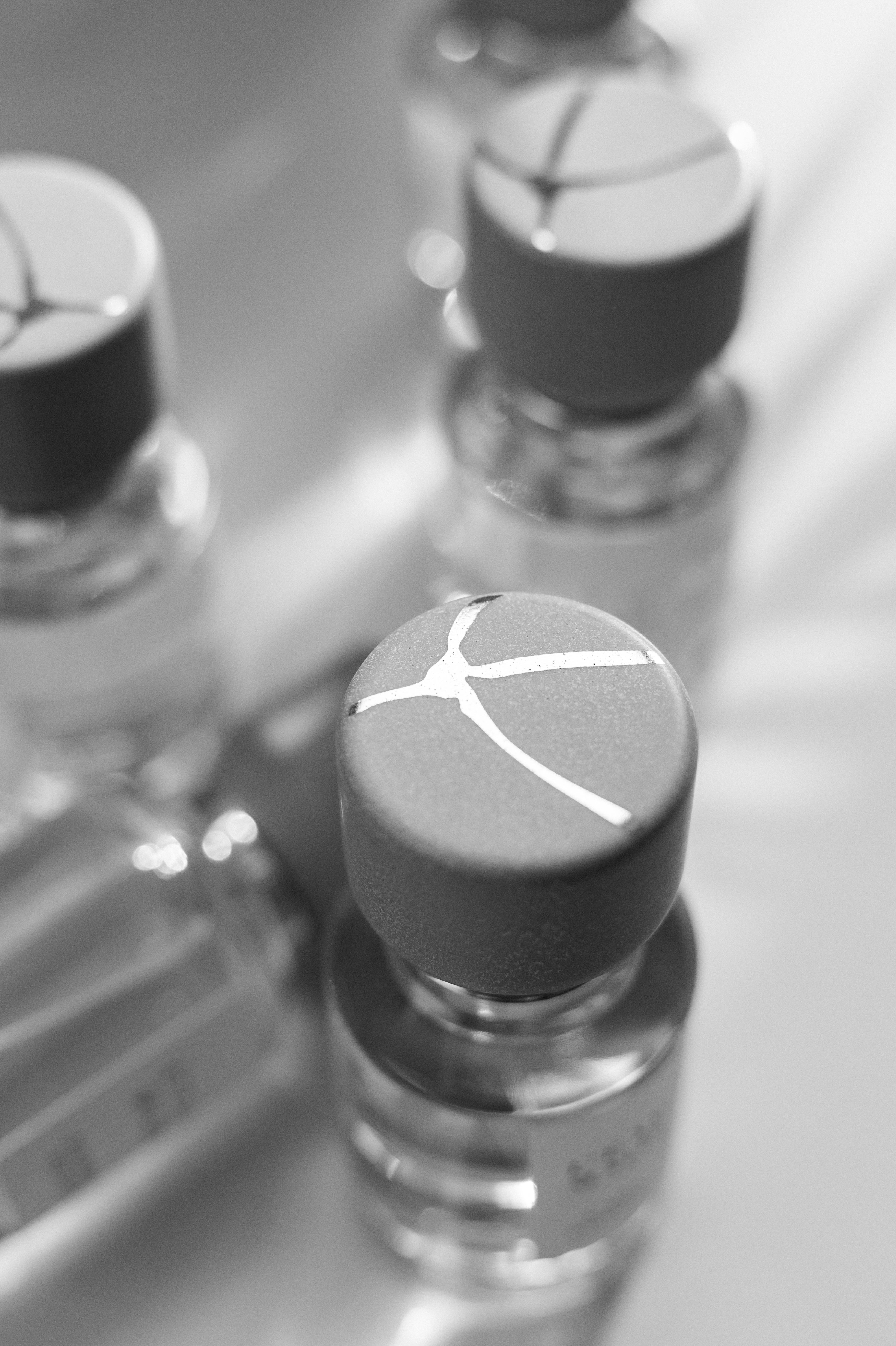 Schwarz-weiß Aufnahme von Parfumflakon der Marke L'eau Maliz mit minimalistischem Logo auf dem Deckel, weitere Flakons unscharf im Hintergrund