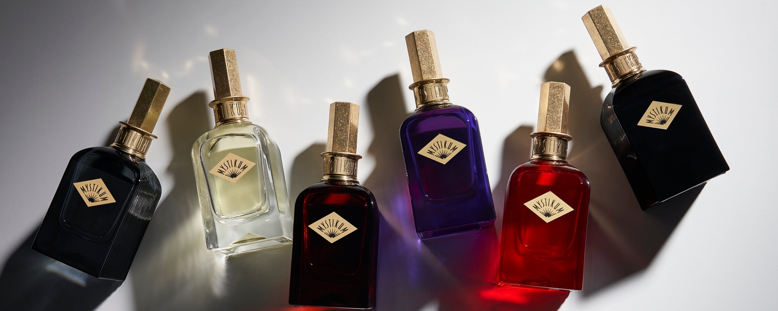 Fünf Parfumflakons von Mystikum in verschiedenen Farben (schwarz, transparent, dunkelrot, lila, rot) mit goldenen Deckeln, dramatisch beleuchtet