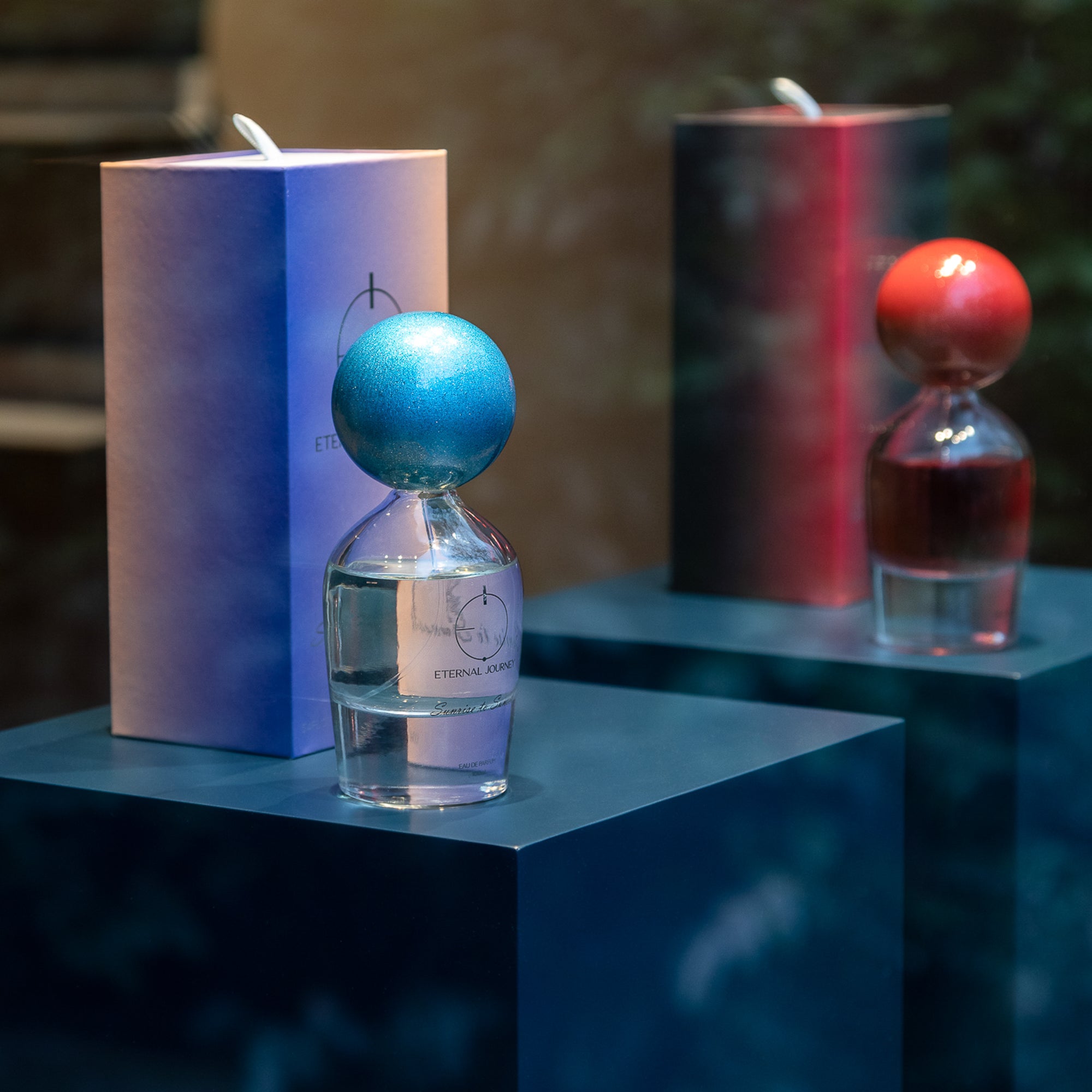 Zwei Parfumflakons mit farbigen Kugelverschlüssen in Blau und Rot auf türkisfarbenen Podesten mit Verpackungen vor unscharfem Hintergrund
