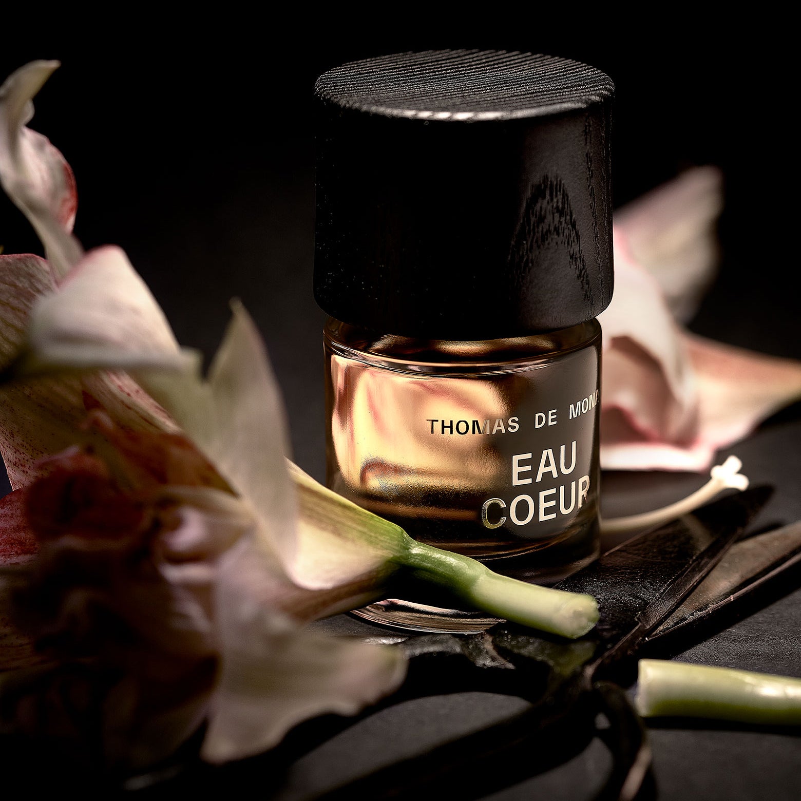 Parfumflakon Eau Coeur mit goldfarbig erscheinender Flüssigkeit und Holzverschluss, arrangiert mit Magnolienblüten und schwarzem Stoff